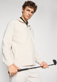 Light beige zip-up jacket made of soft fabric, featuring a black Nike logo, standing collar, and elastic cuffs, with a golf club in hand.