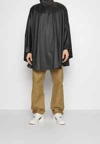 Svart vattentät poncho med hög krage och dragsko, kombinerad med beige byxor och vita sneakers. Slät textur, lös passform.