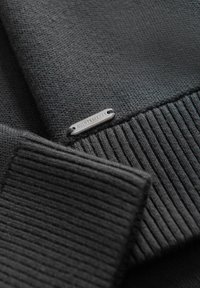 Sort strikket sweater med ribbet manchetter og en glat tekstur. En lille sølvmetal mærke med "QUINTESSENCE" er hæftet på.