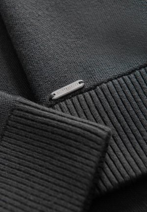 Musta neulepusero, jossa on ribbireunat ja sileä pinta. Pienessä hopeisessa metallitagissa lukee "QUINTESSENCE".