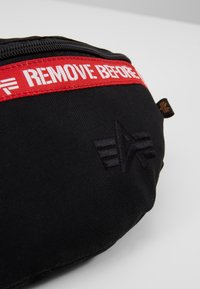 Pochette en tissu noire avec une bande de fermeture éclair rouge étiquetée "REMOVE BEFORE" et un petit logo noir brodé à l'avant.