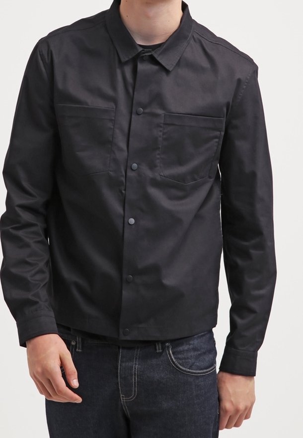 Veste noire boutonnée en tissu résistant. Elle comporte deux poches poitrine, un col, et une coupe courte, portée avec un jean foncé.