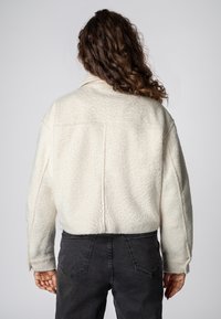 Cream-colored, textured fleece jacket with a collar, slightly cropped fit, and side pockets, paired with dark grey jeans.