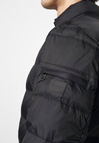 Black puffer jacket with a high collar, featuring a zippered pocket on the sleeve and a textured logo patch on the arm.
