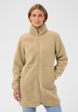Femme portant une veste polaire beige zippée intégralement, avec un col haut et des manches longues, debout, une main dans la poche, sur un fond uni.