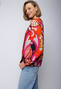 Bunte Bluse mit Blumenmuster in Pink, Rot, Orange und Schwarz, mit langen Ärmeln, elastischen Bündchen und einer lockeren Passform, kombiniert mit hellblauen Jeans.