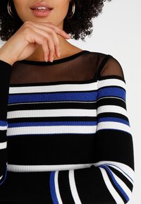 Schwarz-weiß gestreifte Langarmbluse mit blauen Akzenten und einem transparenten Mesh-Ausschnitt. Rippstruktur und taillierte Passform.