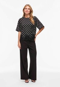 Blouse noire à pois avec manches courtes, finition en dentelle à l'ourlet, associée à un pantalon noir à jambes larges et des sandales. Texture de tissu lisse.