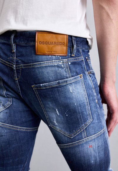 Close-up of blue distressed DSQUARED2 jeans with leather brand patch, worn by a person in a white shirt, showing back pocket and side seam details.