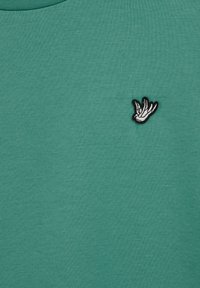 Mėlynos spalvos medvilninė palaidinė su mažesne juoda ir balta siuvinėta paukščio detale kairėje viršutinėje pusėje. Lygus paviršius ir apvalios iškirptės dizainas.