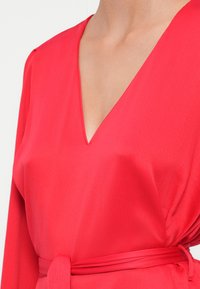Robe portefeuille rouge texturée avec un profond décolleté en V, des manches longues et une ceinture assortie. Le tissu lisse tombe élégamment sur le torse.