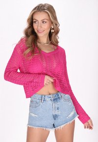 Rosa gehäkelter Pullover mit V-Ausschnitt und langen Ärmeln, kombiniert mit zerrissenen Denim-Shorts mit ausgefransten Kanten.