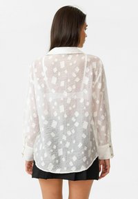 Vrouw met lang haar, gekleed in een doorschijnende witte blouse met vierkante patronen en een zwarte rok, gezien van achteren tegen een witte achtergrond.