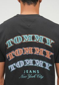 Juodas marškinėliai su daugiaspalviu "TOMMY" tekstu, kuris pateiktas skirtingais šriftais ir dydžiais. Apačioje esantis tekstas "JEANS" ir "New York City" yra mažesniame dydyje.