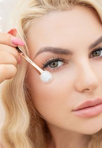 Applicatore rosa dorato con una punta rotonda e bianca in uso sulla zona sotto gli occhi. La modella ha lunghi capelli biondi ondulati e trucco naturale.