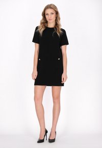 Rochia scurtă, cu mâneci scurte, de culoare neagră are o croială lejeră, două buzunare frontale cu nasturi aurii și un design curat, minimalist. Este purtată cu tocuri negre.