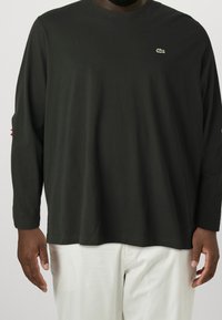 Černé bavlněné tričko s dlouhým rukávem, kulatým výstřihem a výšivkou loga na hrudi. Celý vzhled doplňují bílé kalhoty.