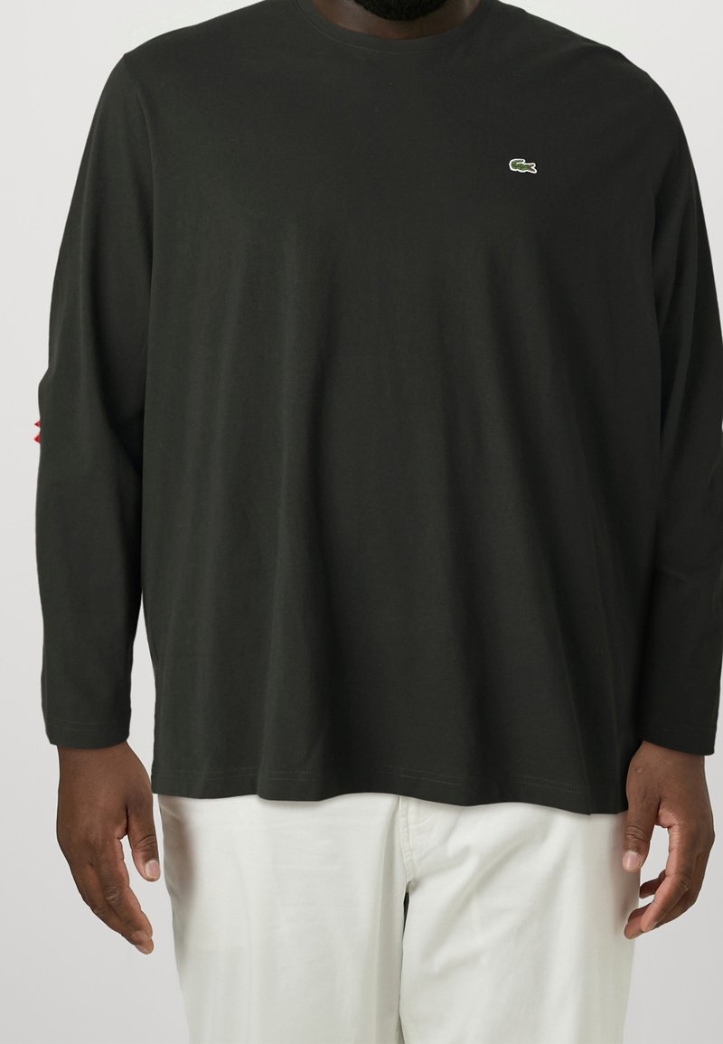 Černé bavlněné tričko s dlouhým rukávem, kulatým výstřihem a výšivkou loga na hrudi. Celý vzhled doplňují bílé kalhoty.