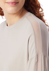 Zbliżenie na osobę ubraną w jasnobeżową bluzkę z transparentną siateczkową wstawką biegnącą wzdłuż ramienia i szwu rękawa.