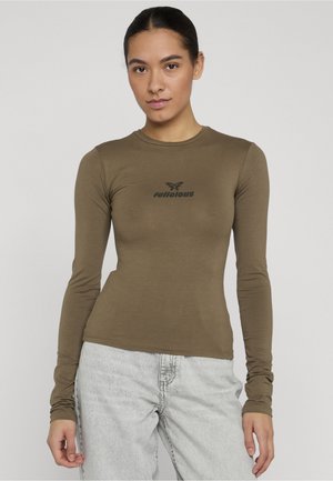 Mladá žena v priliehavom olivovom tričku s dlhým rukávom s nápisom „felicious“ a svetlosivých džínsoch stojaca pred jednoduchým pozadím.