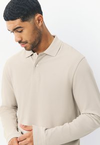 Beige pitkähihaisen poolopaidan, jossa on hienovarainen logo, napituslista ja teksturoitu kangas. Siinä on klassinen kaulus ja istuvat kalvosimet.