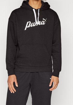 Personne portant un sweat à capuche noir Puma avec des cordons blancs et un pantalon Puma noir assorti, debout devant un fond uni.