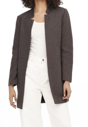 Femme portant un blazer marron foncé ouvert sur un haut blanc et un pantalon blanc taille haute, debout avec les bras détendus.