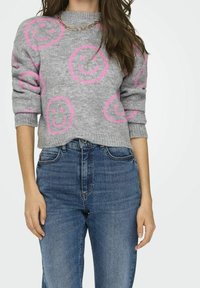 Szary sweter dzianinowy z różowymi wzorami uśmiechniętych buziek, z podwiniętymi rękawami i okrągłym dekoltem, w zestawie z niebieskimi dżinsami z wysokim stanem. Złoty naszyjnik z łańcuszka.