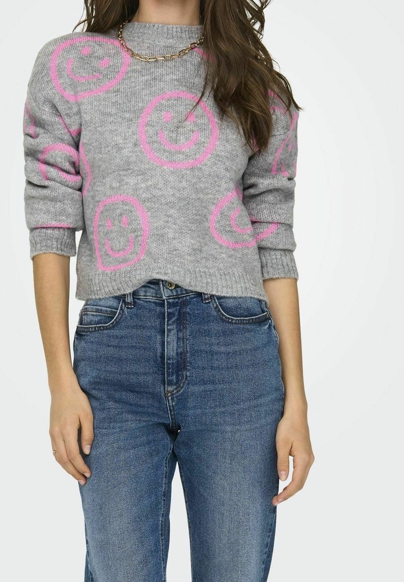 Szary sweter dzianinowy z różowymi wzorami uśmiechniętych buziek, z podwiniętymi rękawami i okrągłym dekoltem, w zestawie z niebieskimi dżinsami z wysokim stanem. Złoty naszyjnik z łańcuszka.