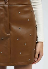 Jupe en simili cuir marron avec de petites clous en argent, portée avec un haut à manches longues côtelé blanc et une main visible reposant sur le côté.