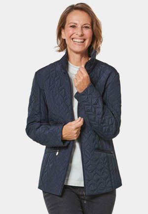 Marineblauwe gewatteerde jas met een staande kraag, twee ritszakken en een gestructureerd ruitpatroon. Draagt over een lichtgekleurde shirt.