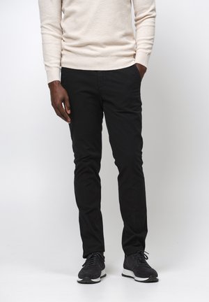 Zwarte katoenen broek in een slanke pasvorm, met een vlakke voorkant en standaardzakken, gecombineerd met een lichtbeige trui en zwarte sportschoenen.