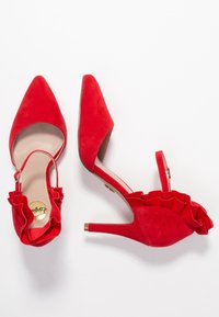Červené suede stiletto boty s špičatou špičkou a volánkovým řemínkem na kotníku. Obsahují zlatý akcent na podpatku. Hladké vnitřní podšívání.