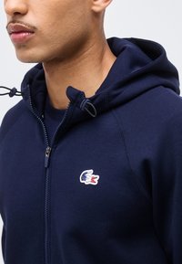 Mann iført marineblå hettejakke med glidelås og en tre-farget krokodillelogo på brystet samt justerbare snorer til hetten.