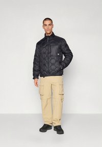 Sort dunjakke med sekskantet quiltning, lynlås og høj krave, kombineret med beige cargobukser. Modellen står på en neutral baggrund.