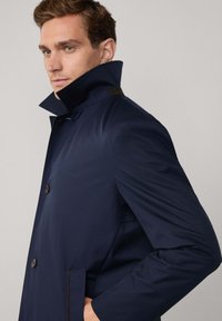 Cappotto blu navy con colletto alto, chiusura a un bottone e tasche laterali. Tessuto liscio, leggermente lucido con una vestibilità su misura.