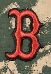 Embroidered letter "B" in red outlined with black on camouflage fabric featuring green and beige patterns, textured surface.