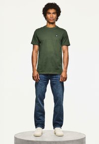 Hombre con cabello rizado vistiendo una camiseta verde, jeans azules y zapatillas blancas, de pie sobre una plataforma redonda contra un fondo liso.