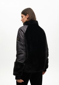 Black padded jacket with faux fur body and collar, quilted sleeves, and large cuffs. Features a gold logo on the left sleeve.