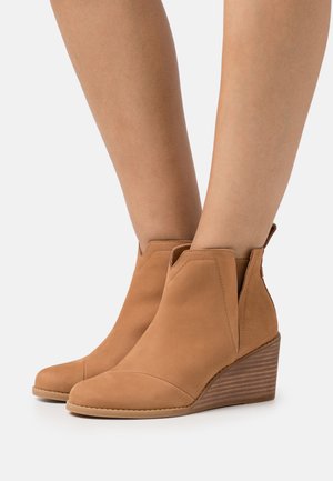 Tan suede wedge ankle boots with a rounded toe, V-shaped cutouts on the sides, and a striped textured wedge heel.