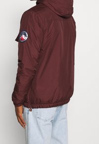 Marjanpunainen hupullinen takki, joka on tehty nailonista, ja siinä on kiristysnyöri helmassa sekä pyöreä logokoriste vasemmassa hihaisessa. Sileä pinta, rento malli.