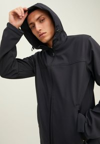 Chaqueta de softshell negra con capucha, que presenta un cierre frontal con cremallera, bolsillos laterales y una textura suave. Diseño ajustado con detalles cosidos.