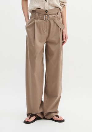 Kobieta w beżowych spodniach o wysokim stanie i szerokich nogawkach z pasującym paskiem, w czarnych sandałach typu thong i lekkim dzianinowym topie.