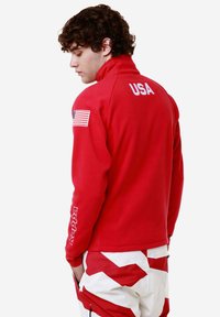 Giacca in pile rossa con colletto alto, "USA" stampato sulla schiena e una toppa con la bandiera americana sulla manica. Abbinata a pantaloni bianchi e rossi a fantasia.