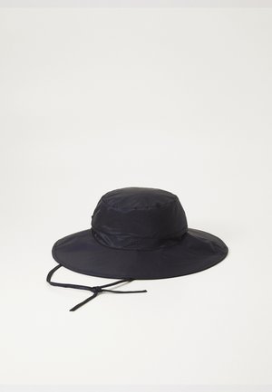Chapeau seau bleu marine en tissu léger, doté d'un large bord et d'une sangle de menton réglable. Surface texturée avec de subtils plis.