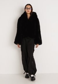 Juoda dirbtinio kailio striukė su gobtuvu, laisvo silueto ir ilgomis rankovėmis. Derinama su juodomis krovinių kelnėmis ir masyvia juoda avalyne.