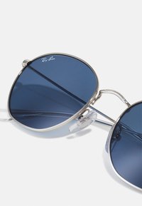 Occhiali da sole rotondi con lenti blu scuro, montatura in metallo argentato e nasioli trasparenti. Presenta il logo Ray-Ban sulla lente.