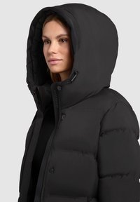 Chaqueta acolchada negra con una gran capucha, textura acolchada, botones a presión y apertura con cremallera. Tela suave con un diseño estilizado.