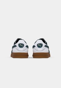 Pár bílých a tmavě zelených tenisek Puma s hnědými podrážkami z pohledu zezadu na jednobarevném bílém pozadí.