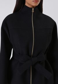 Cappotto di lana nero con colletto alto, chiusura frontale a zip e cintura in vita con fiocco. Il tessuto ha una texture morbida e un design strutturato.